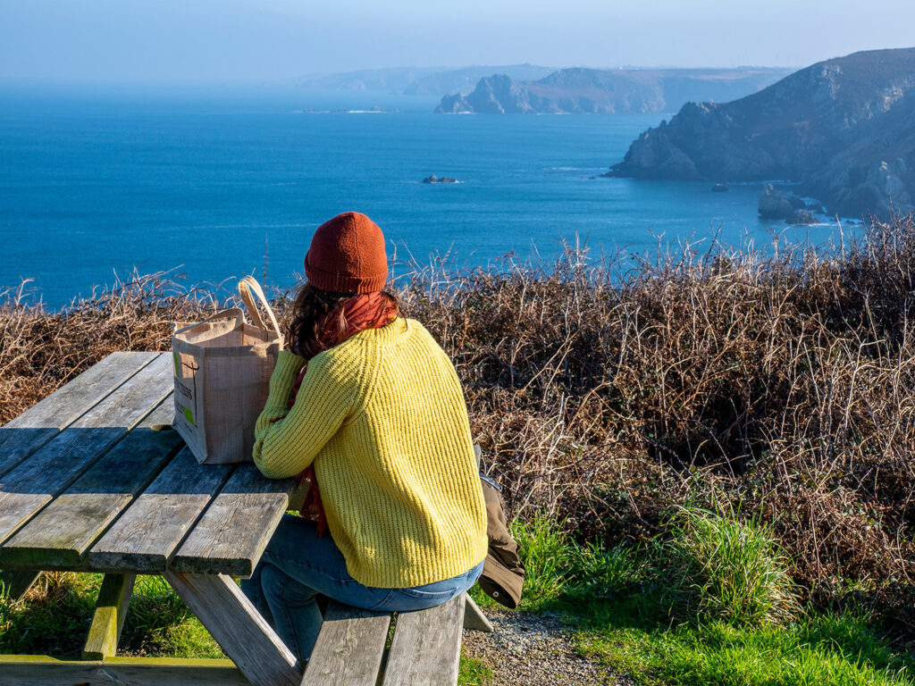 dame i gul genser og rød lue på picknikbenk med utsikt over havet
