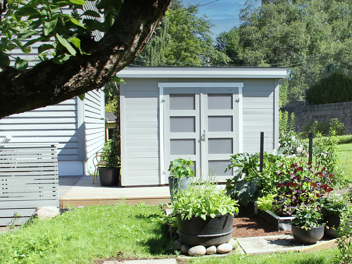 Grå bod i hage på platting med kjøkkenhage i front. Info i bildet på størrelse og fordeler. Foto.